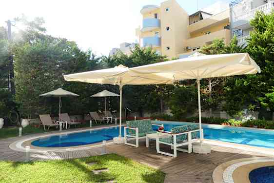 Ulkoallas, jossa aurinkotuoleja ja varjoja, ympäröitynä puilla ja rakennuksilla. (Tekoälyn luoma teksti)