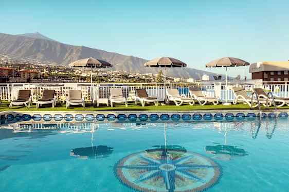 Uima-allasalue, jossa aurinkotuoleja ja aurinkovarjoja, näkymä vuorille ja rakennuksiin. (Tekoälyn luoma teksti)