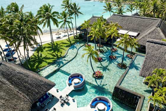 Useita olkikattoisia taloja uima-altaan vieressä, taustalla palmuja ja hiekkaranta. (Tekoälyn luoma teksti)