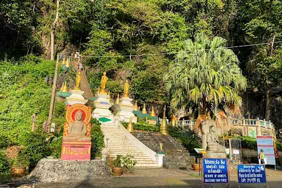 Kuva portaat, jotka johtavat useisiin kultaisiin buddhalaisiin patsaisiin ja rakenteisiin vehreyden ympäröimänä. (Tekoälyn luoma teksti)