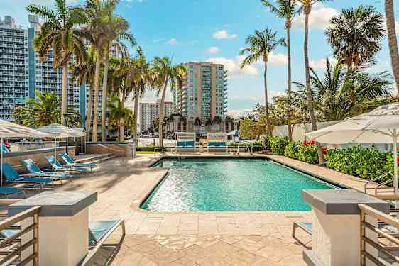 Uima-allas ympäröitynä aurinkotuoleilla ja aurinkovarjoilla, taustalla näkyy korkeita rakennuksia ja palmupuita. (Tekoälyn luoma teksti)