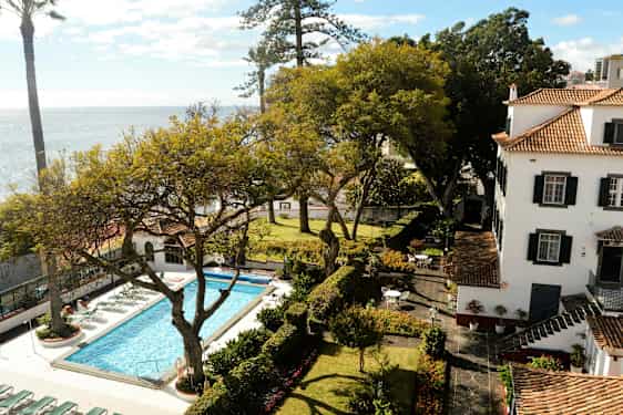 Kuva puutarhasta, missä on uima-allas, ja se on puiden ympäröimä, taustalla rakennus. (Tekoälyn luoma teksti)