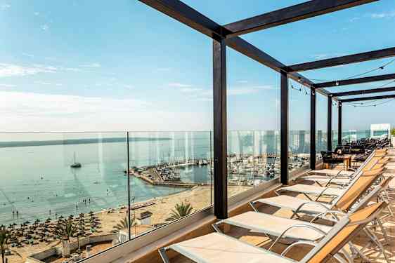 Aurinkotuoleja asetettu lasikaiteen eteen, näkymä rannalle ja satamaan. (Tekoälyn luoma teksti)