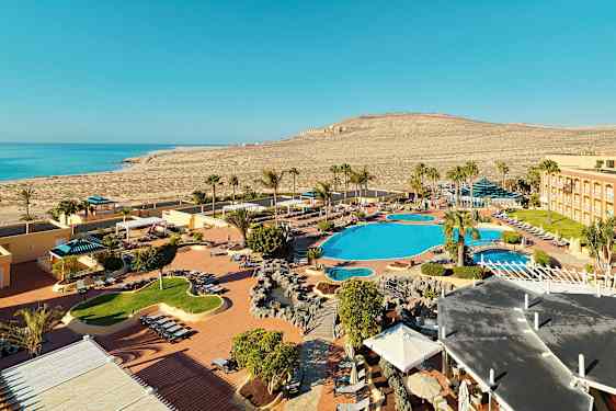 Kuvassa on lomakohde rannalla, jossa on uima-allasalueita, aurinkotuoleja ja palmuja. Taustalla näkyy merta ja ympäröivää kuivaa maisemaa. (Tekoälyn luoma teksti)