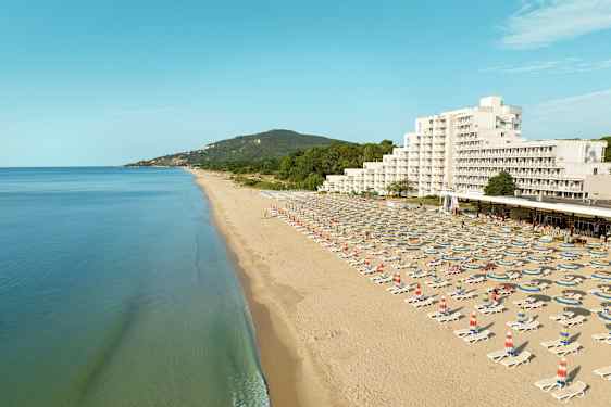 Pitkä hiekkaranta aurinkotuoleineen ja varjoineen, taustalla hotelli ja vihreitä kukkuloita. (Tekoälyn luoma teksti)