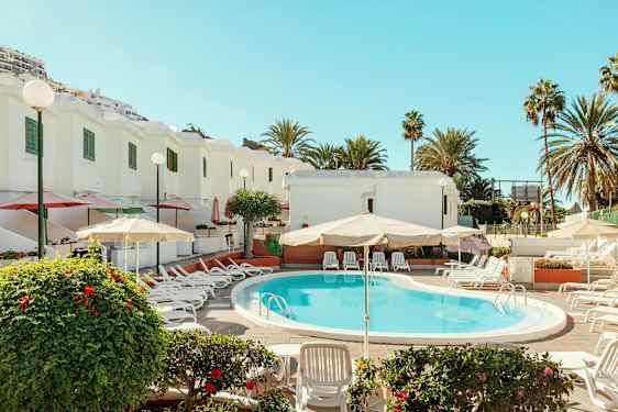 Ulkouima-allas, jonka ympärillä aurinkotuoleja ja sateenvarjoja, valkoisten rakennusten vieressä, palmuja ympärillä. (Tekoälyn luoma teksti)