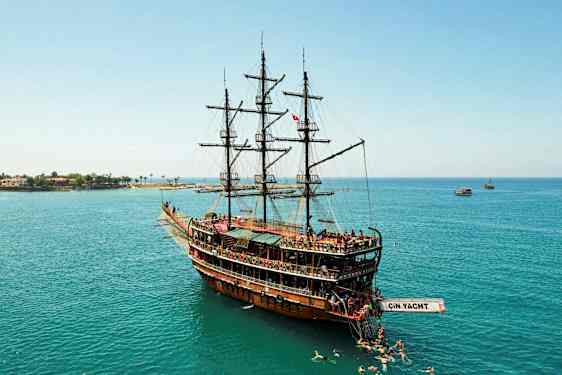 Suuri purjevene puusta, joka kelluu rauhallisilla vesillä uimareiden ympäröimänä, taustalla rannikkoa. (Tekoälyn luoma teksti)