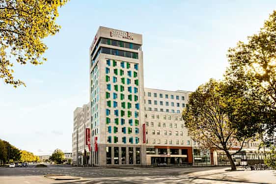 Moderni hotellirakennus, jossa on valkoinen julkisivu ja värikkäät ikkunat, ympärillä teitä ja puita. (Tekoälyn luoma teksti)