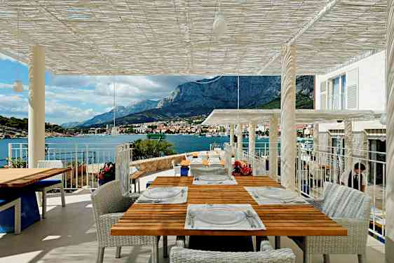 Terassi, jolta avautuu näkymä vuorille ja merelle, useat pöydät ovat katettu vieraita varten. (Tekoälyn luoma teksti)