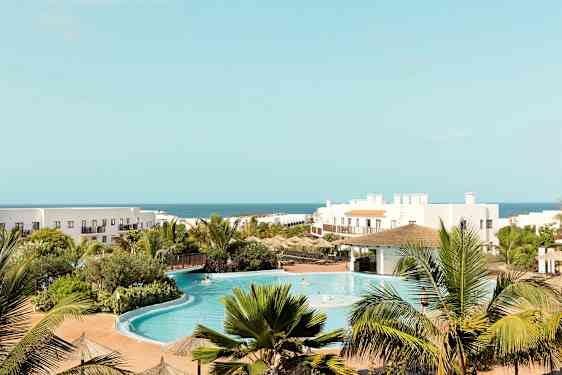 Hotellikompleksi, jossa on uima-allas, vehreää ympäristöä ja taustalla meri. (Tekoälyn luoma teksti)