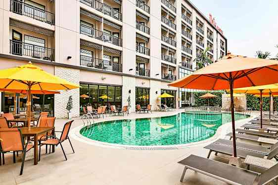 Hotellin uima-allas, jota ympäröivät aurinkotuolit ja varjot, taustalla rakennus. (Tekoälyn luoma teksti)