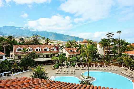 Hotellialue, jossa on uima-allas, jota ympäröivät aurinkotuolit ja vehreä ympäristö, taustalla vuoria. (Tekoälyn luoma teksti)