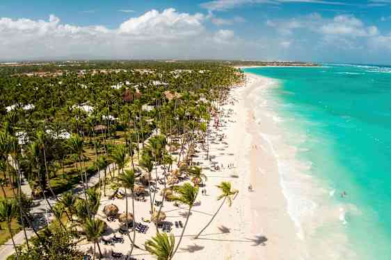 Valkoinen hiekkaranta ja turkoosi vesi sekä paljon palmuja. Rannan kävijät ja rantatuolit näkyvät pitkin rantaa. (Tekoälyn luoma teksti)