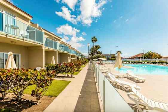 Kuva hotellialueesta, jossa on uima-allas, aurinkotuoleja ja vehreyttä. Rakennuksessa on parvekkeita. (Tekoälyn luoma teksti)