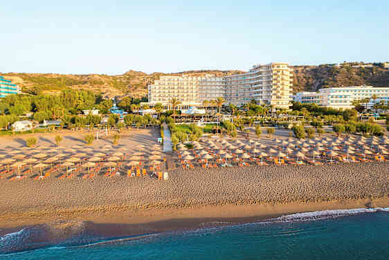 Ranta aurinkovarjoilla ja aurinkotuoleilla, taustalla hotelli ja vihreitä alueita. (Tekoälyn luoma teksti)