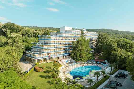 Kuva hotellista, jossa on useita kerroksia ja parvekkeita, ympäröitynä vehreällä kasvillisuudella. Allas on etualalla aurinkotuoleineen. (Tekoälyn luoma teksti)