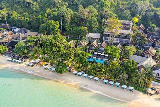 Ilmakuva rannasta, jossa on aurinkotuoleja ja -varjoja sekä rakennuksia vehreyden keskellä. (Tekoälyn luoma teksti)