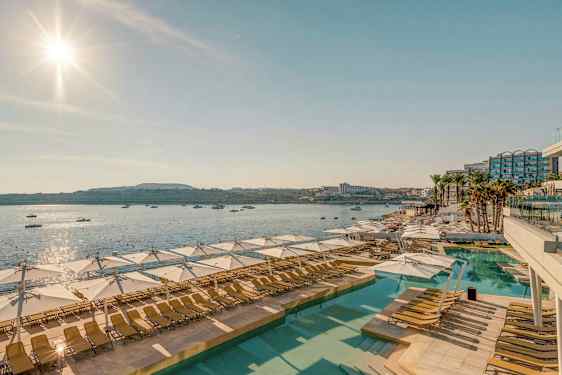 Näkymä aurinkoiselle uima-allasalueelle, jossa on aurinkotuoleja ja aurinkovarjoja, taustalla meri ja veneet. (Tekoälyn luoma teksti)