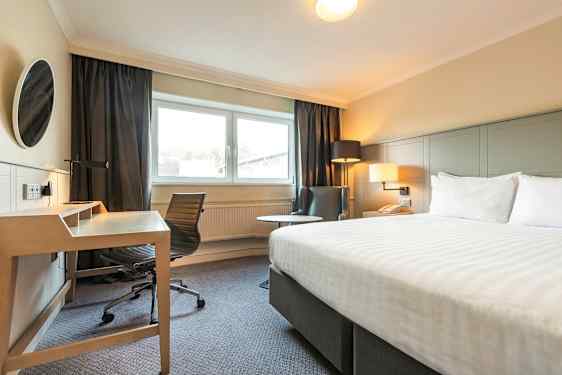 Kuva hotellihuoneesta, jossa on sänky, työpöytä ja tuoli sekä nojatuoli ikkunan vieressä. (Tekoälyn luoma teksti)