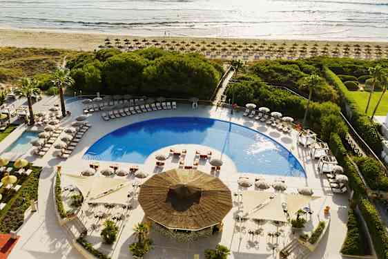 Näkymä uima-allasalueelle, jossa aurinkotuoleja ja aurinkovarjoja, ympärillä vehreyttä lähellä rantaa. (Tekoälyn luoma teksti)