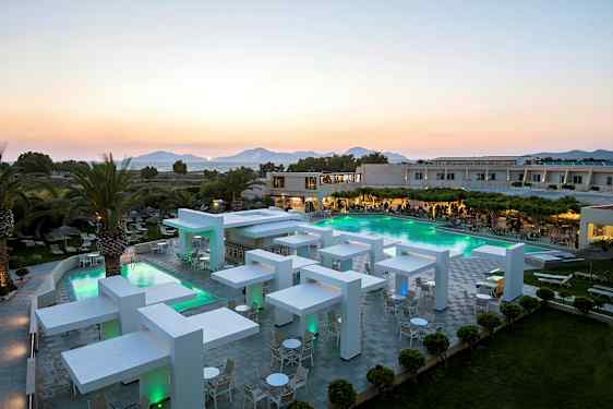 Moderni hotellialue, jossa uima-allas ja iltavalaistus, ympäröity vehreydellä ja palmupuilla, taustalla vuoret. (Tekoälyn luoma teksti)