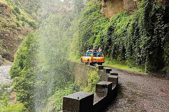 Kelta-oranssi jeep kahden hengen joukolla tiellä vesiputouksen kupeessa vihreässä laaksossa. (Tekoälyn luoma teksti)
