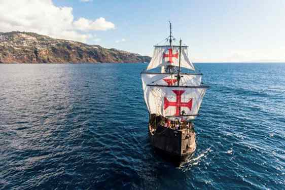 Purjelaiva, jonka suuret valkoiset purjeet on koristeltu punaisilla risteillä, purjehtii sinisen meren yllä lähellä mäkistä rannikkoa. (Tekoälyn luoma teksti)