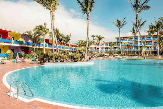 Suuri uima-allas palmuilla ympäröitynä, taustalla värikkäitä hotellirakennuksia. (Tekoälyn luoma teksti)