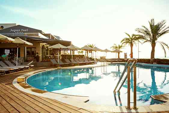 Ulkouima-allas, jonka ympärillä aurinkotuoleja ja varjoja. Taustalla näkyy rakennus, jossa lukee 'Aegean Pearl'. (Tekoälyn luoma teksti)