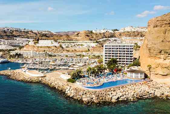 Kuva rannikkokaupungista, jossa satama, rinteessä olevia rakennuksia ja hotelli, jossa uima-allas. (Tekoälyn luoma teksti)