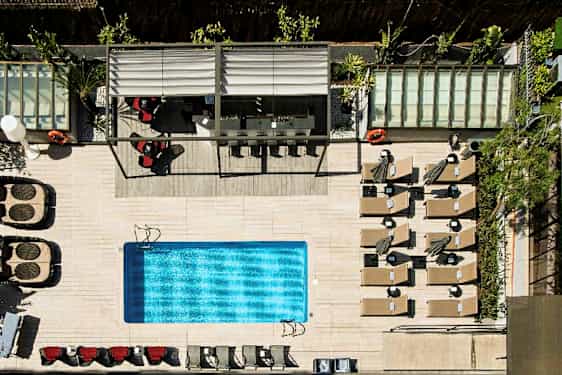 Lintuperspektiivistä otettu kuva ulkouima-altaasta, jota ympäröivät aurinkotuolit, aurinkovarjot ja katettu istuma-alue. (Tekoälyn luoma teksti)