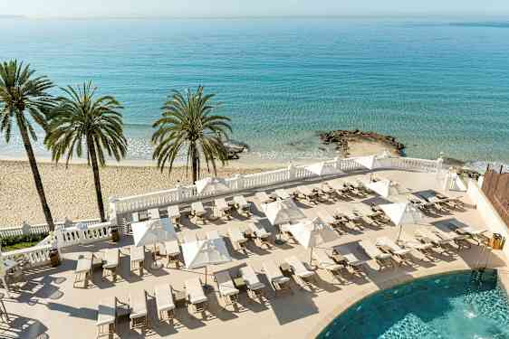 Ranta, jossa palmuja, aurinkotuoleja ja varjoja, sinisen meren äärellä. (Tekoälyn luoma teksti)