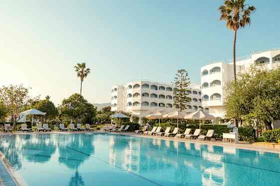 Hotellirakennus uima-altaan kanssa, aurinkotuoleja ja aurinkovarjoja etualalla. (Tekoälyn luoma teksti)