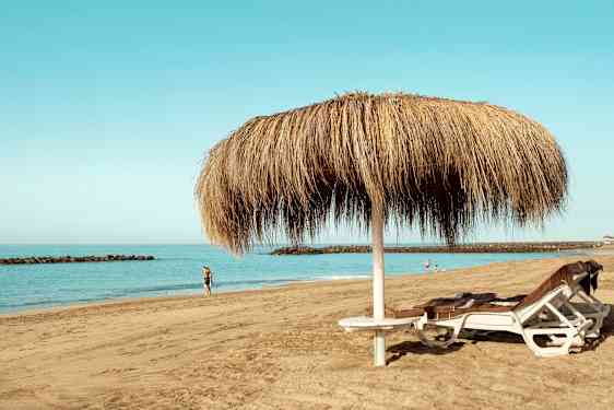 Kuva hiekkarannasta, jossa kaksi aurinkotuolia olkivarjon alla ja ihmisiä kävelemässä veden äärellä. (Tekoälyn luoma teksti)