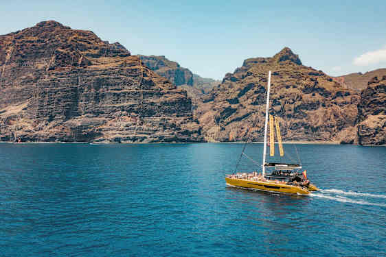 Katamaraani purjehtii lähellä korkeita kallioita, kirkkaan sininen vesi etualalla. (Tekoälyn luoma teksti)