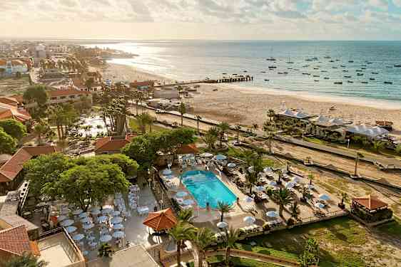 Ilmakuva rannasta, jossa aurinkotuoleja ja varjoja lähellä uima-allasta, vihreyden ympäröimänä. (Tekoälyn luoma teksti)