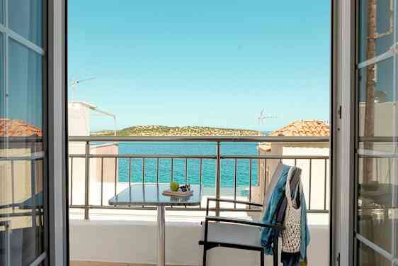 Näkymä huoneesta ovien läpi parvekkeelle, pöytä, tuoli ja vaatteita, sekä meri ja maisema taustalla. (Tekoälyn luoma teksti)