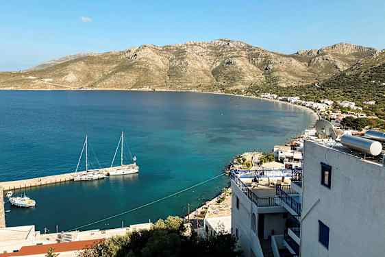 Näkymä rannikolle, jossa pieniä rakennuksia ja vuoria taustalla. Veneet ovat kiinnitettyinä laituriin kirkkaassa vedessä. (Tekoälyn luoma teksti)