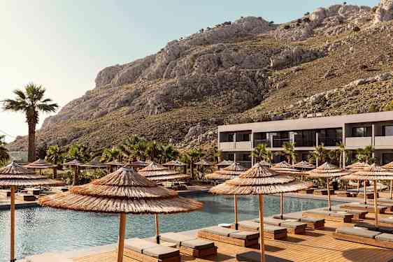 Nykyaikainen rakennus parvekkeineen sijaitsee uima-altaan vieressä, jota ympäröivät aurinkotuolit ja aurinkovarjot. Taustalla näkyy kallioita ja palmupuita. (Tekoälyn luoma teksti)