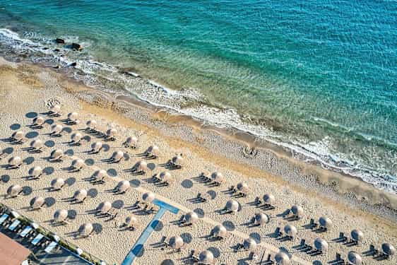 Ilmakuva hiekkarannasta, jossa aurinkotuoleja ja aurinkovarjoja, taustalla rantaviiva ja sininen meri. (Tekoälyn luoma teksti)