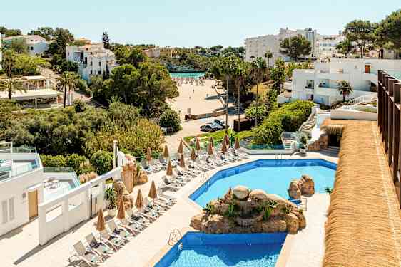 Hotellialue, jossa uima-allas, aurinkotuoleja ja aurinkovarjoja, taustalla näkyy ranta ja valkoiseksi maalattuja rakennuksia. (Tekoälyn luoma teksti)