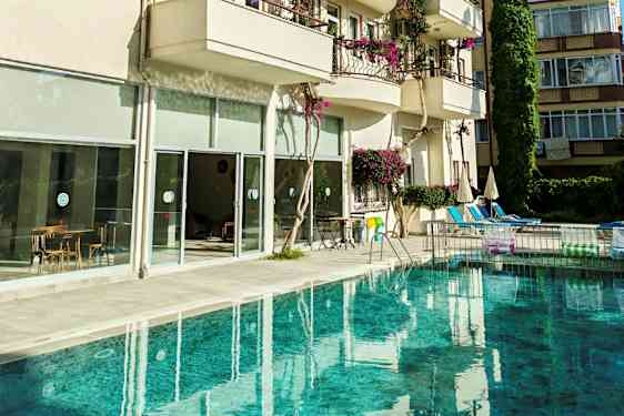 Uima-allas rakennuksen edessä, jossa on parvekkeita ja suuria lasi-ikkunoita. Kuvassa näkyy myös tuoleja, pöytiä ja aurinkovarjoja altaan ympärillä. (Tekoälyn luoma teksti)