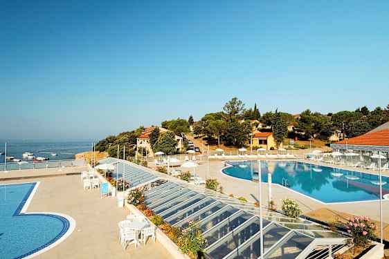 Ulkoallas, jota ympäröi terassi, jossa on aurinkotuoleja ja aurinkovarjoja. Taustalla näkyvät meri sekä rakennukset ja kasvillisuus. (Tekoälyn luoma teksti)