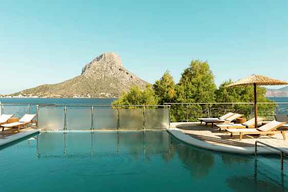 Ulkoallas, jossa aurinkotuoleja ja varjo, sijaitsee lähellä merta, taustalla kukkula. (Tekoälyn luoma teksti)