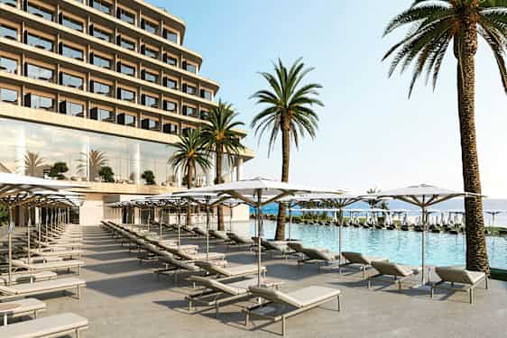 Kuvassa hotellin uima-allas, jonka etualalla on aurinkotuoleja ja varjostimia, ja taustalla näkyy suuria palmuja ja hotellin julkisivu. (Tekoälyn luoma teksti)