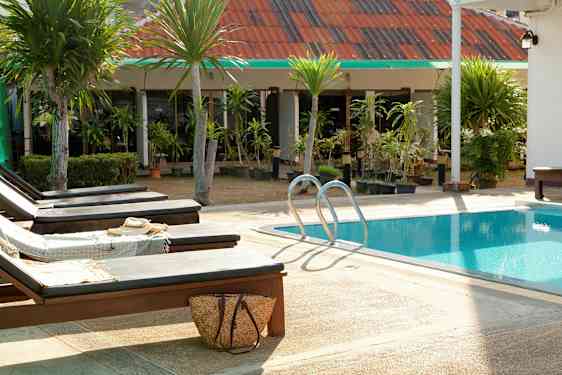 Aurinkotuoleja uima-altaan äärellä, taustalla vihertäviä puita ja rakennus. (Tekoälyn luoma teksti)