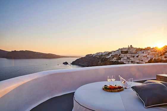 Näköala auringonlaskuun meren rannalla, jossa valkoisia rakennuksia kallion laella ja terassi pöytineen, ruokaineen ja juomineen etualalla. (Tekoälyn luoma teksti)