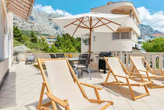 Aurinkotuoleja ja aurinkovarjo terassilla, josta on näkymä vuorelle ja ympäröiviin rakennuksiin. (Tekoälyn luoma teksti)