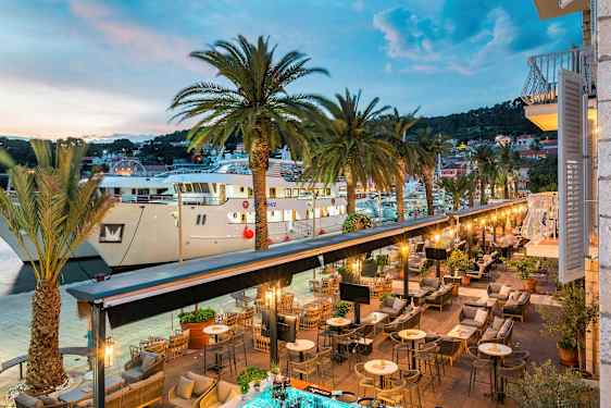 Näkymä ulkoilmaravintolasta satamaan, jossa on palmupuita ja suuria veneitä. (Tekoälyn luoma teksti)