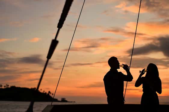 Kaksi henkilöä siluettina, jotka pitävät laseja auringonlaskun aikaan, taustalla meri ja pieni saari. (Tekoälyn luoma teksti)